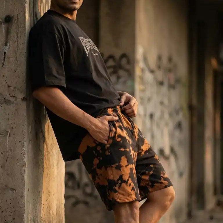 Male model leaning casually against a concrete pillar in an urban setting, styling black and brown bleach wash shorts with a black graphic tee and chunky white sneakers.