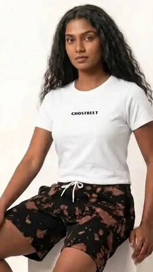 Female model sitting on a white display block, wearing Ghostreet black and brown tie-die sweat shorts paired with a white Ghostreet graphic t-shirt and white sneakers. — Photo 3