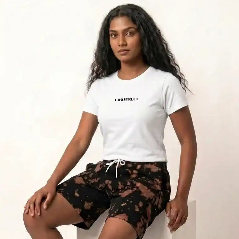 Female model sitting on a white display block, wearing Ghostreet black and brown tie-die sweat shorts paired with a white Ghostreet graphic t-shirt and white sneakers.