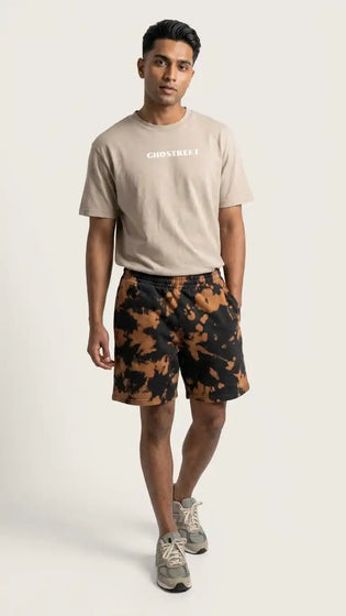 Male model walking towards the camera, wearing relaxed fit black and rust tie-dye sweat shorts, a beige Ghostreet t-shirt, and grey sneakers. — Photo 1