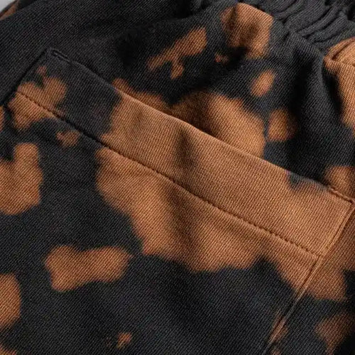 Close-up fabric detail of black and rust-brown bleach wash sweat shorts, focusing on the stitching and the back patch pocket.